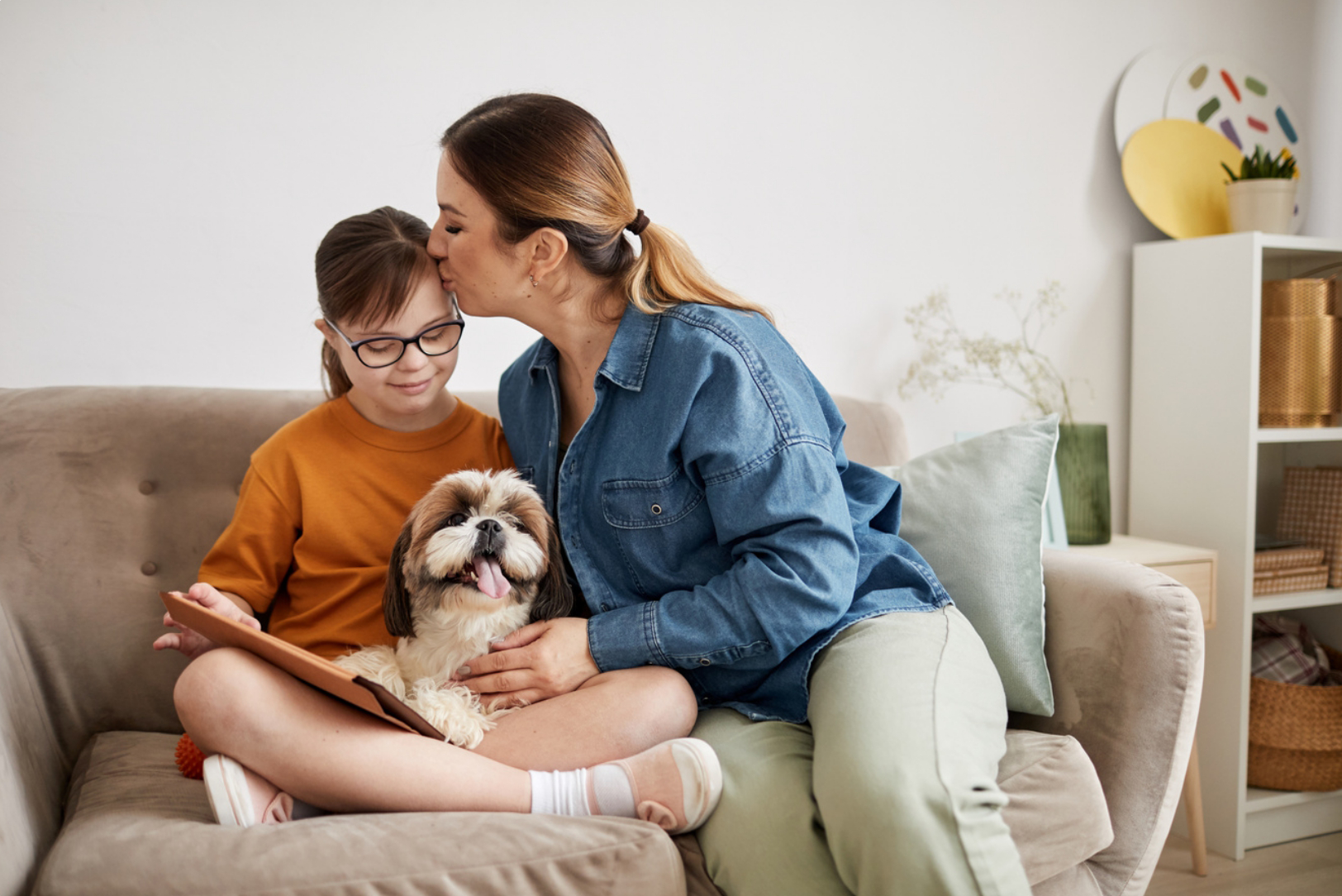 Mother kissing daughter on the head on a sofa with a dog, representing caring for family
